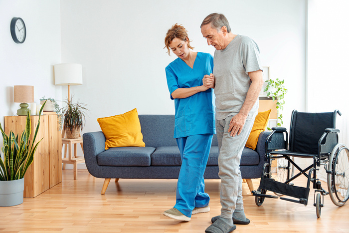 Young caregiver assisting senior man in wheelchair walking indoors - Home health care service